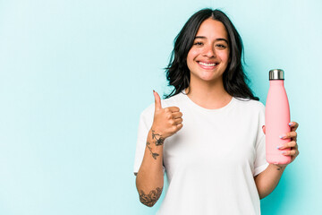 Young hispanic woman holding thermo isolated on blue background smiling and raising thumb up