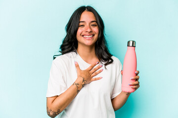 Young hispanic woman holding thermo isolated on blue background laughs out loudly keeping hand on...