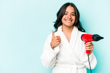 Young hispanic woman holding dryer isolated on blue background smiling and raising thumb up
