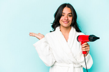 Young hispanic woman holding dryer isolated on blue background showing a copy space on a palm and...