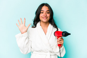 Young hispanic woman holding dryer isolated on blue background smiling cheerful showing number five with fingers.