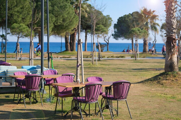 Empty tables and colored chairs in a coastal cafe restaurant in city park near the trees on the embankment against the backdrop of the sea and people