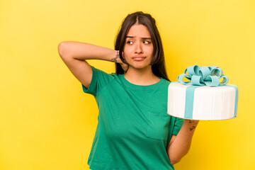 Young hispanic woman holding birthday cake isolated on yellow background touching back of head, thinking and making a choice.