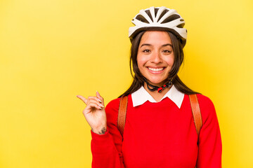 Young student hispanic woman wearing a bike helmet isolated on yellow background smiling and pointing aside, showing something at blank space.