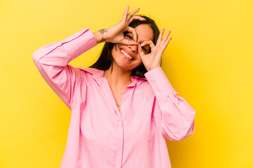 Young hispanic woman isolated on yellow background showing okay sign over eyes