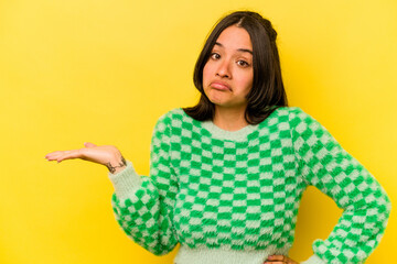 Young hispanic woman isolated on yellow background showing a copy space on a palm and holding another hand on waist.