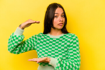 Young hispanic woman isolated on yellow background holding something with both hands, product presentation.