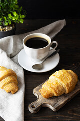 Crispy croissant on a light brown wooden plank. Black coffee in a white cup in the background. Light napkin on a wooden table