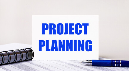 On a light gray background, a notebook, a blue pen and a sheet of paper with the text PROJECT PLANNING. Business concept