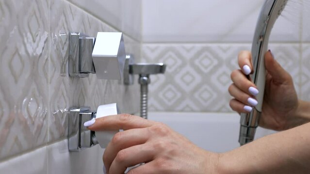 Woman turns a steel faucet in a modern bathroom adjusting the supply of warm water for washing. Square shaped metal tap female hand turns to adjust the temperature of the water from the shower head