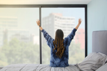 Rear view of a woman stretching her arms after waking up and getting out of her bed, looking out...