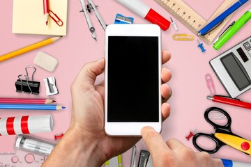 Girl holding a smartphone with a blank screen. Pink background with school supplies, accessories,