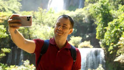 Fototapeta premium Happy social media content maker. Smiling influencer talk on video, shoot blog on mobile phone outdoors. Adult man blogger, vlogger recording selfie movie on his smartphone at nature, look at camera.