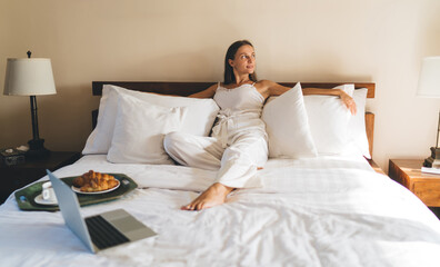 Skilled female lying at comfortable hotel bed with modern laptop computer and looking away - thinking about service dayspa, carefree woman with netbook resting during weekend morning in home interior