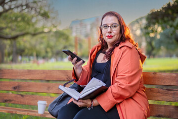 Woman sitting on a bench in a park checking her agenda and cell phone while having a coffee