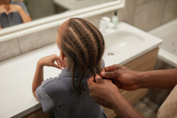 Close up of caring black father braiding daughters hair in bathroom and helping get ready in morning