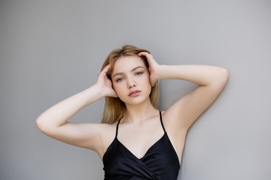 Stylish young woman touching hair and looking at camera on grey background.