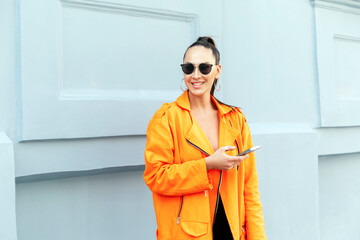 Young stylish girl on a city street with a phone. Beautiful brunette in a bright orange jacket and sunglasses. Modern life of youth.