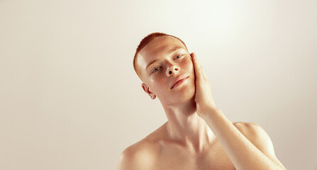 Portrait of young red-haired man with freckles touching cheeks, posing isolated over grey studio background. Shaving lotion