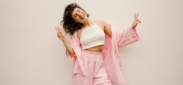 Funny young caucasian girl smiles eyes closed, showing peace gesture on white background. Brunette with short haircut wears top, shirt and pants. Mood, lifestyle, concept.