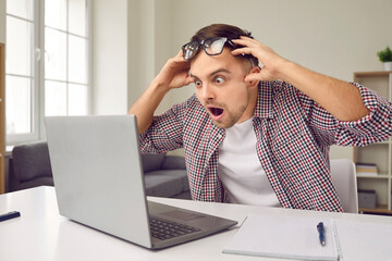 Excited stunned man with shocked expression reads email on his laptop screen with favorable proposition. Guy is sitting at table in front of laptop and grabs his head and can not believe his luck.