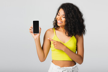 happy african american woman pointing at smartphone with blank screen isolated on grey.