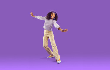 Funny African American teenager in stylish eccentric clothes isolated on purple studio background dancing. Smiling biracial teen girl have fun make dancer moves. Hobby and entertainment concept.