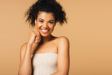 happy young african american woman with bare shoulders looking at camera isolated on beige.