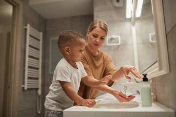 Portrait of young mother helping cute toddler boy brushing teeth in bathroom, copy space
