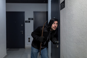 A young male robber in dark clothes stands carefully and quietly at the door of the apartment in...