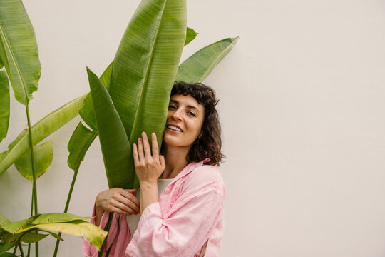 Cute young caucasian girl looking at camera hugging palm leaves on white background. Brunette with short haircut wears pink shirt. Mood, lifestyle, concept.