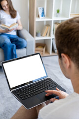 Digital life. Computer mockup. Home routine. Unrecognizable man working laptop with blank screen while defocused woman reading book in light room interior.