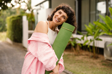 Happy young caucasian girl with yoga mat posing at camera outdoors. Brunette with short haircut wears pink shirt. Good mood concept