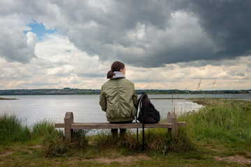 Fototapeta premium Rear view of a woman sitting on a bench alone looking at the view across the water, stormy sky.