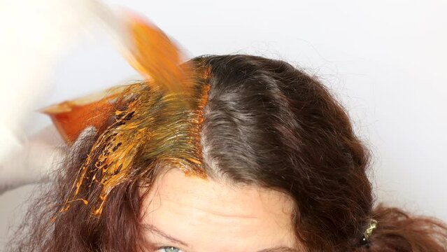 Cropped shot of hairdresser wearing white gloves, applying orange hair dye from bowl on long dark brown curly hair with rat-tail brush, dyeing hair roots on white background. Hair colouring. Close-up.