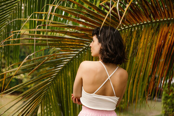 Attractive young caucasian woman posing with her back to camera in front of palm tree on street. Brunette with short haircut looks away and wears white top. Camping concept