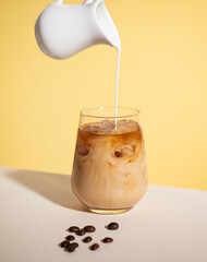 Iced coffee with poured cream in a glass with ice cubes and grains on a yellow background. The...