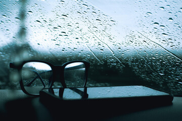 Lentes de leer junto a un celular en prime plano frente a una ventana con lluvia con paisaje de...