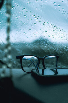 Lentes de leer junto a un celular en prime plano frente a una ventana con lluvia con paisaje de playa desenfocada en d&iacute;a nublado