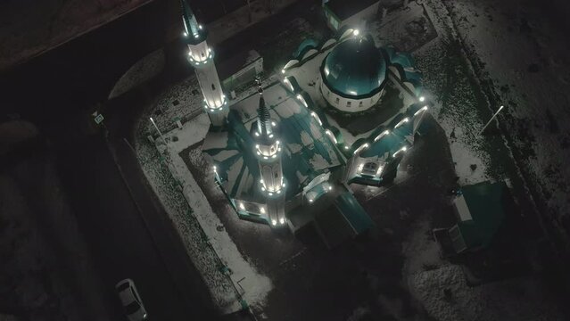 Historical Muslim mosque building with powerful light equipment on snowy street of evening town in winter bird eye view