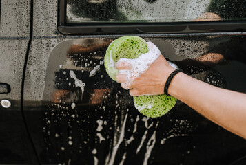 A male professional worker, a washer, with a green washcloth in his hands, washes the black surface of a car door with white soapy foam. Photography, shampoo pattern concept, copy space.