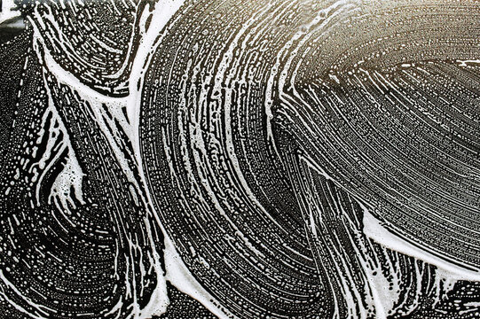 Background, texture of the black surface of a metal car door in white soapy foam after washing. Photo of shampoo streaks.