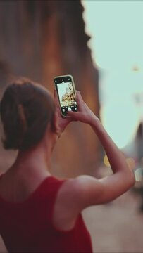 VERTICAL VIDEO: Close-up, girl takes pictures of downtown mobile phone. Closeup of young woman shoots the cityscape on smartphone in the evening time. Back view