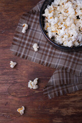 Heap of popcorn in black bowl on wooden rustic background. Some popcorns on fabric and table. Vertical shot, top view