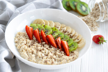Bowl of oatmeal with fresh fruits, strawberry, banana, kiwi and linseeds with kitchen towel, jar with oats and fruits on background. Recipe of healthy breakfast, clean eating.