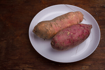 Orange, purple raw sweet potatoes, yam on white plate, wooden rustic table. Farm harvest, organic vegetables, shop local vegan food