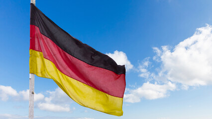 The official flag of Germany waving on a blue sky background. Horizontal banner design, with the German flag hanging on a sunny background with white clouds. Deutschland flag wide banner
