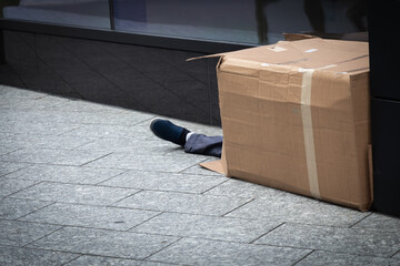 un homme sans domicile fixe vivant dans la rue dort dans un carton. Sa jambe dépasse du carton....