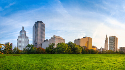 Panoramic Downtown Hartford Cityscape over the green park with the blue sky and white clouds at sunrise. Beautiful tranquil morning landscape of modern downtown buildings in natural settings.