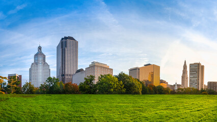 Obraz premium Panoramic Downtown Hartford Cityscape over the green park with the blue sky and white clouds at sunrise. Beautiful tranquil morning landscape of modern downtown buildings in natural settings.